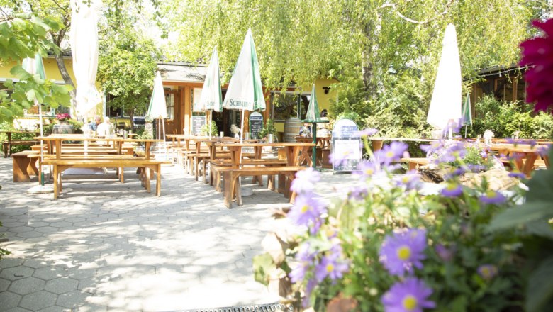 Guest garden, © Ben Leitner A sunny guest garden with wooden benches and tables, surrounded by green trees and colorful flowers.