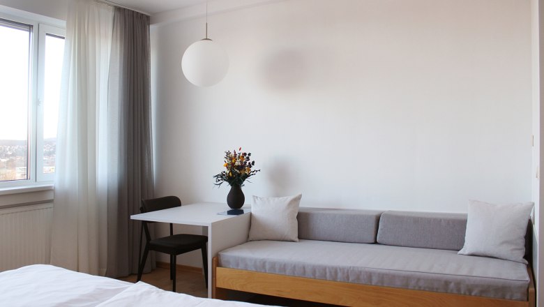 Gästezimmer, © Stadthotel Hollabrunn Modernes Gästezimmer mit Sofa, Tisch, Stuhl und Fenster mit Vorhängen.