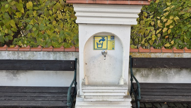 Trinkbrunnen, © Weinviertel Tourismus GmbH Ein kleiner Brunnen mit Ziegeldach und Wasserhahn, flankiert von zwei Holzbänken auf einem gepflasterten Platz.