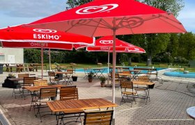 Freibad Niederkreuzstetten, © Gemeinde Kreuzstetten Ein Freibad mit roten Sonnenschirmen und leeren Holztischen auf einer gepflasterten Terrasse.