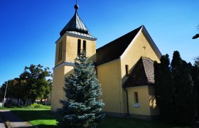 Hetzmannsdorf Kapelle, © Weinstraße Weinviertel Gelbe Kapelle mit Turm und Kreuz, umgeben von Bäumen, unter blauem Himmel.