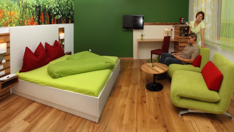 Guest room, © Familie Bohrn A modern guest room with a green bed, sofa and wooden floor. There are two people in the room, one sitting on the sofa, the other standing next to it.