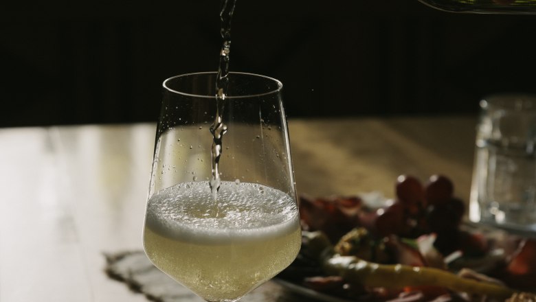 Wein einschenken, © Weinviertel Tourismus / Sophie Menegaldo Ein Glas Weißwein wird auf einem Tisch eingeschenkt, im Hintergrund ein Teller mit Snacks.