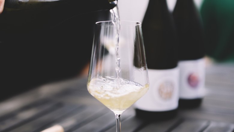 Wein einschenken, © Niederösterreich Werbung / Mara Hohla Weißwein wird in ein Glas auf einem Tisch eingeschenkt, mit Flaschen im Hintergrund.
