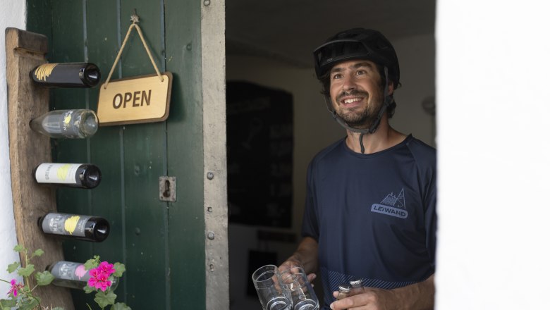 Rest stop in the Öhlbergkellergasse, © Weinviertel Tourismus GmbH / Frühmann A man with a bicycle helmet stands smiling in an open doorway, holding glasses and bottles. An "Open" sign hangs on the door.