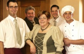 Familie Schweinberger, © Fam. Schweinberger Fünf Personen in traditioneller Kleidung und Kochuniform posieren lächelnd in einem Raum.