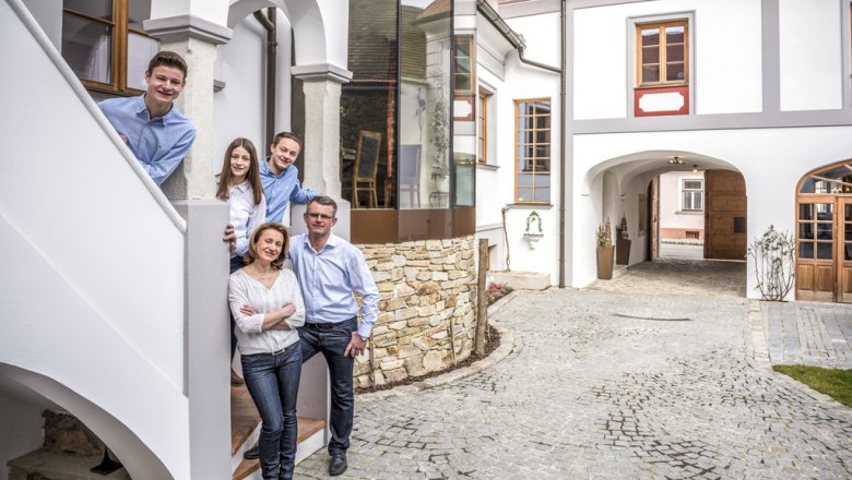 Am Weingut, © Mediadesign Podolsky Eine Familie steht lächelnd auf einer Treppe vor einem Weingut mit gepflastertem Hof.