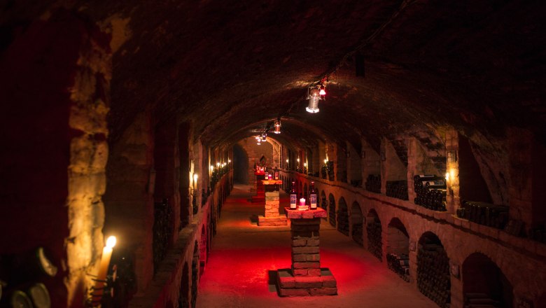 Kellerlabyrinth, © Umschaid Ein stimmungsvoll beleuchteter Weinkeller mit Flaschen auf Steinsäulen.