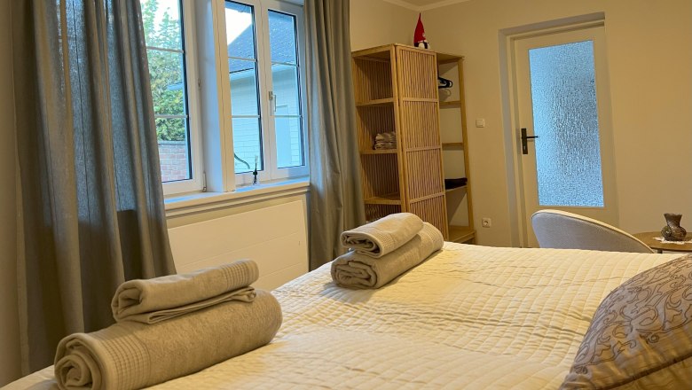 Bedroom, © Waltraud Forstner A cozy bedroom with a bed with towels on it, a window with curtains and a shelf with decorations.