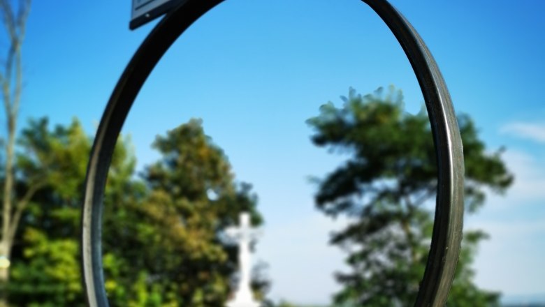 Schlüsselerlebnis Zeißlkreuz, © Weinstraße Weinviertel Ein Schild mit der Aufschrift 'Seefeld-Kadolz' vor einem blauen Himmel, im Hintergrund ein weißes Kreuz und Bäume.