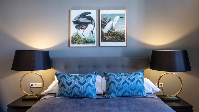Doppelzimmer, © Astrid Bartl Doppelzimmer mit grauem Bett, blauen Kissen, zwei Nachttischlampen und Vogelbildern an der Wand.