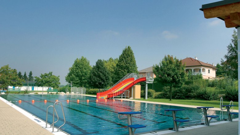 Freibad, © Gemeinde Unterstinkenbrunn Ein Freibad mit einem Schwimmbecken, einer roten Rutsche und Sprungbrettern. Im Hintergrund sind Bäume und ein Haus zu sehen.