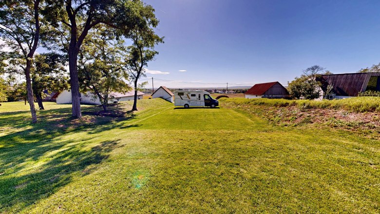 Spritzerrast – Campingstellplatz mit Jause und Panoramablick, © Bernhard Raab Wohnmobil auf einem grünen Campingplatz mit Bäumen und Gebäuden im Hintergrund.