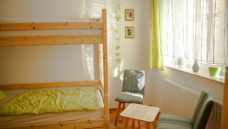 Stockbett, © Michael Sokolar Ein helles Zimmer mit einem Stockbett, zwei Stühlen, einem kleinen Tisch und einem Fenster mit Vorhängen.
