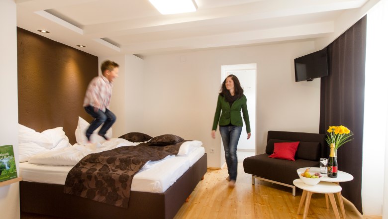 Weingut Sonnenhügel, © Astrid Bartl Ein Kind springt auf einem Bett in einem modernen Hotelzimmer, während eine Frau lächelnd zusieht. Ein Tisch mit Blumen und Wein steht daneben.