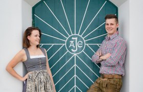 Stephanie und Lukas, © Christopher Anker Ein Mann und eine Frau in traditioneller Kleidung vor einer grünen Tür mit geometrischem Muster.