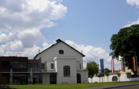 Konzerthaus Weinviertel, © Marktgemeinde Ziersdorf Ein modernes Konzerthaus mit weißen Wänden und großen Fenstern, umgeben von Flaggen und Bäumen, unter einem bewölkten Himmel.