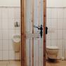 Zwei Toilettenräume mit Holztür, links ein Urinal und Waschbecken, rechts eine Toilette.