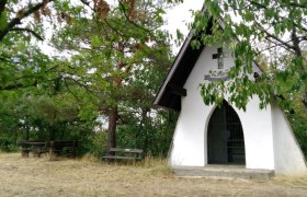 Kleine wei&szlig;e Kapelle im Wald mit Holzb&auml;nken davor.