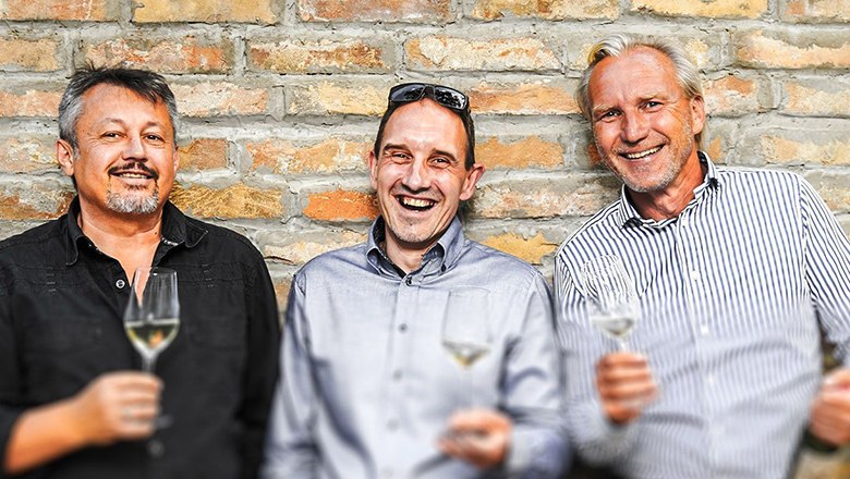 Drei Männer stehen vor einer Ziegelwand und halten Weingläser in der Hand.