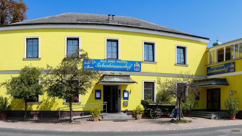 Gelbes Gebäude mit Restaurant und Hotel, umgeben von Bäumen und Straße.