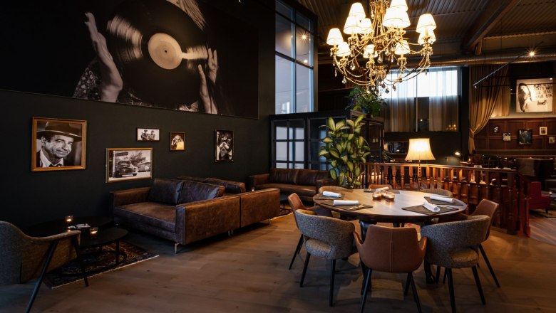 Ein stilvoll eingerichteter Raum mit Ledersofas, einem runden Tisch und einem großen Kronleuchter. An der Wand hängen Schwarz-Weiß-Fotografien.