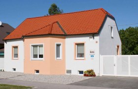 Ein kleines, einstöckiges Haus mit orangefarbenem Dach und weißer Fassade unter blauem Himmel.