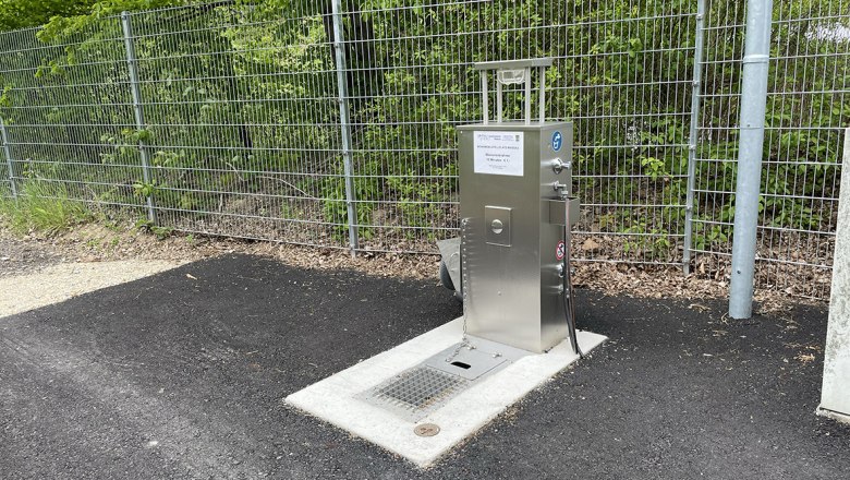 Metallene Wasserversorgungsstation neben einem Zaun auf Asphalt.