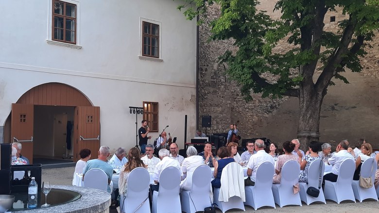 Menschen sitzen an einem langen Tisch im Freien, umgeben von historischen Gebäuden und Bäumen.