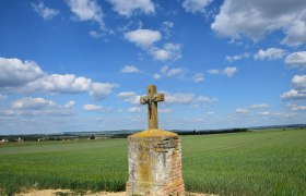 Ein steinernes Kreuz mit Christusfigur steht auf einem Feld unter blauem Himmel mit Wolken.
