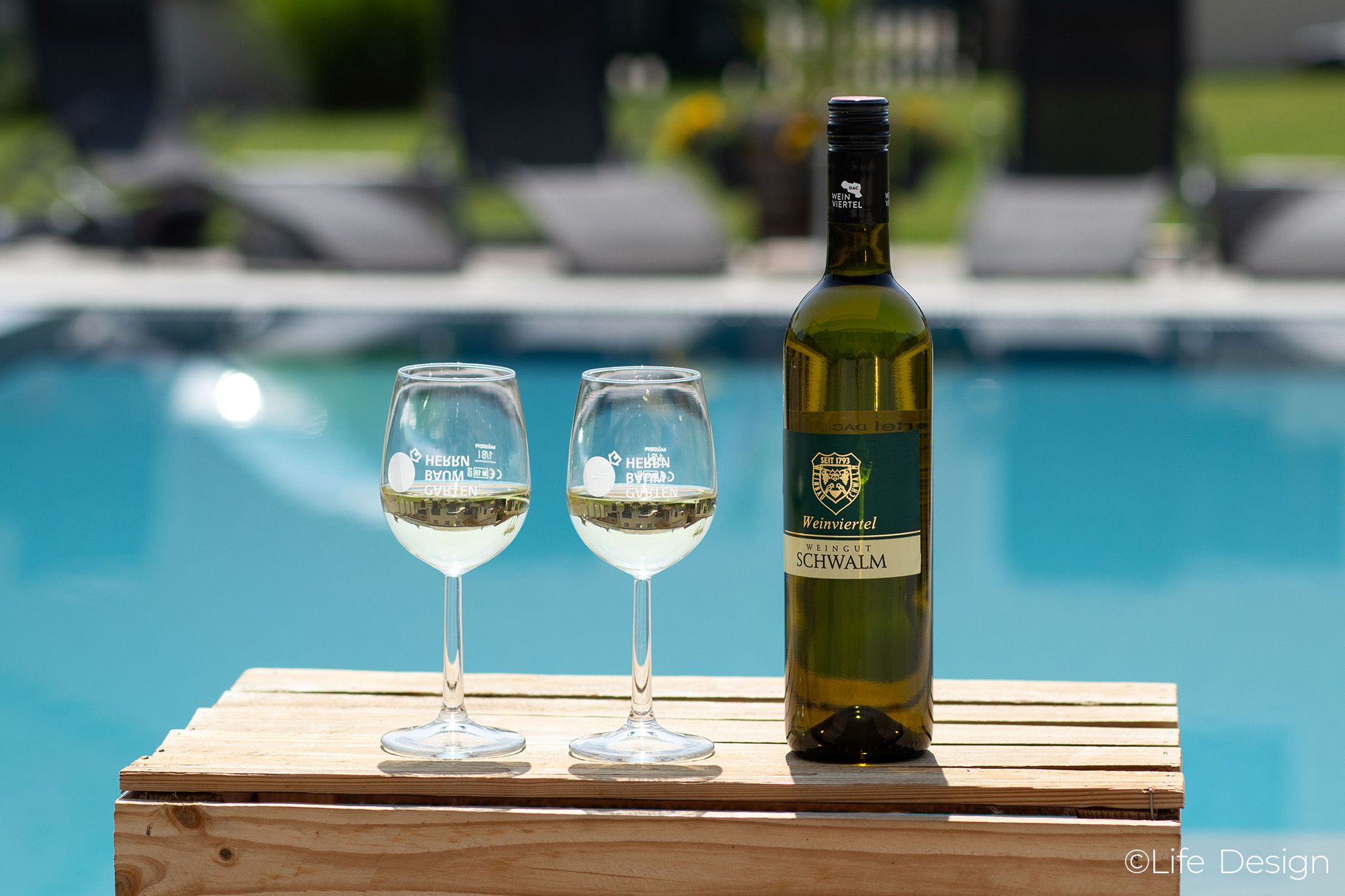 Eine Flasche Wein und zwei Weingläser auf einem Holztisch vor einem Pool.
