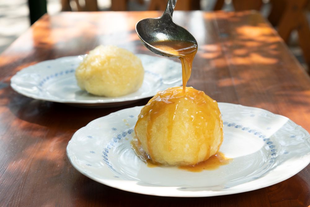 Ein Knödel auf einem Teller wird mit Soße übergossen, ein weiterer Knödel im Hintergrund.