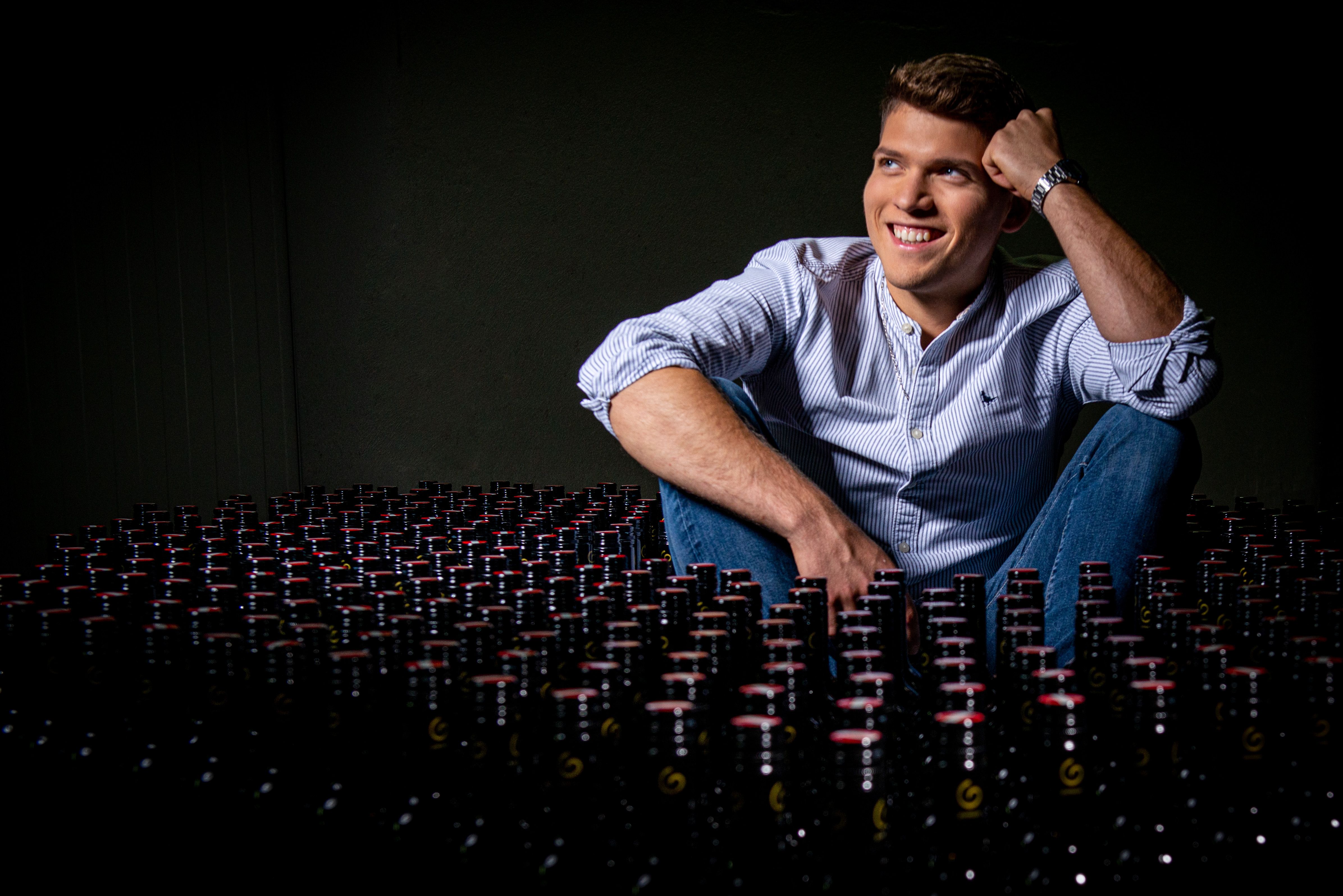A man sits smiling in a wine cellar, surrounded by many bottles of wine.