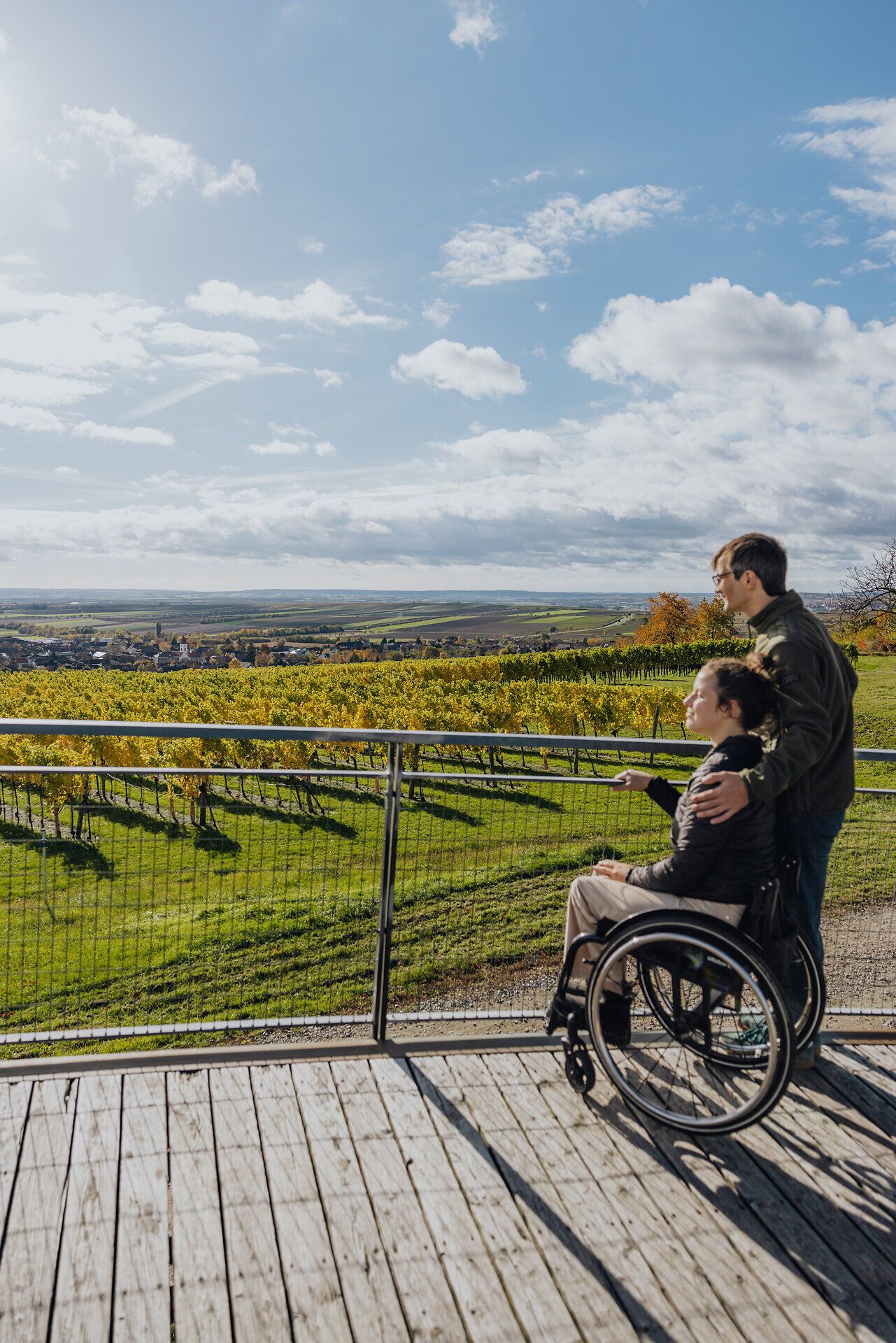 Eine Frau im Rollstuhl und ein Mann stehen auf einer Aussichtsplattform und blicken auf die herbstlich gefärbten Weingärten und die Weite der Weinviertler Landschaft.