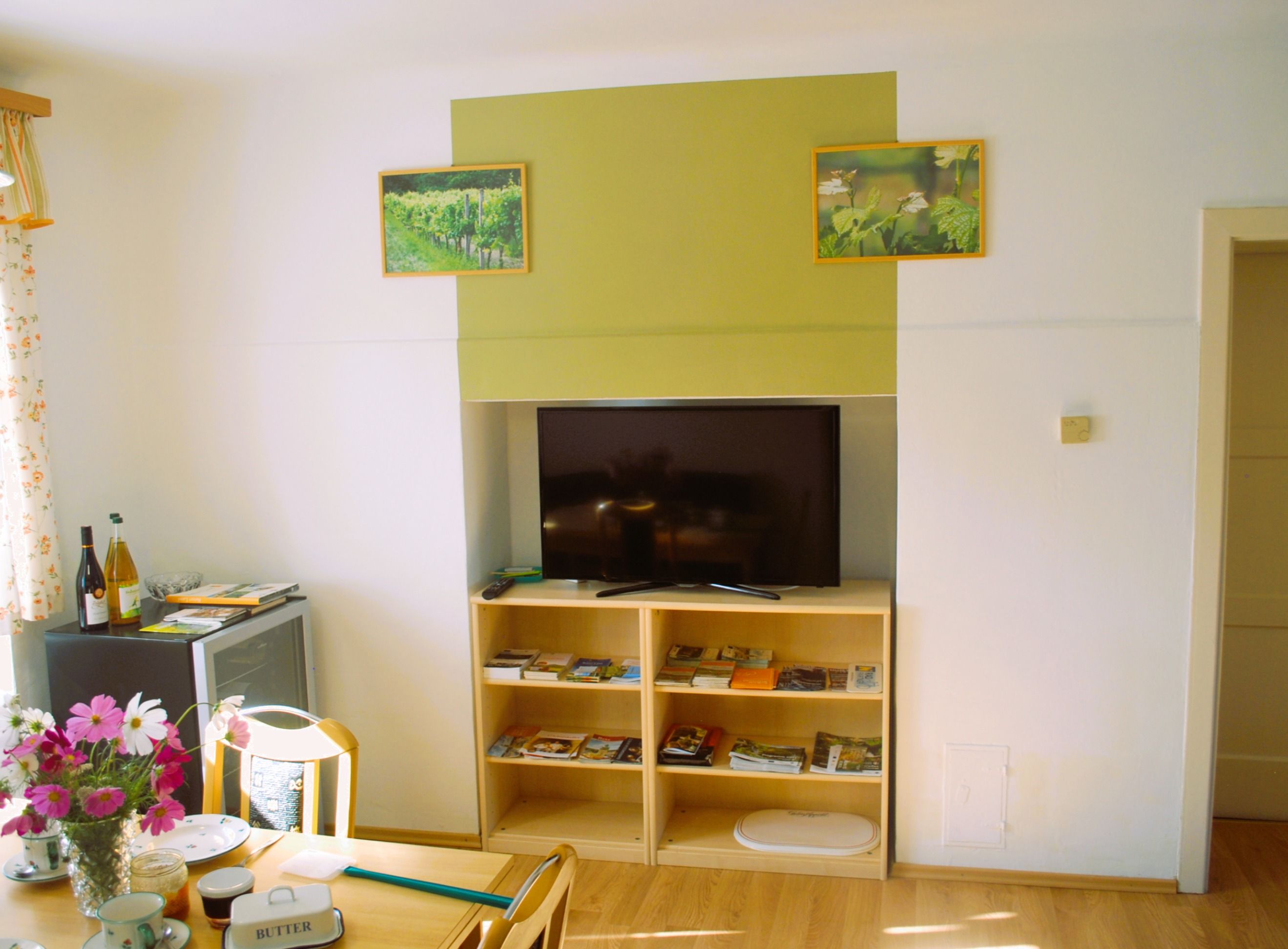 Ein Wohnzimmer mit einem Fernseher auf einem Regal, umgeben von Broschüren. An der Wand hängen zwei Bilder von Weinbergen. Ein Tisch mit Blumen und Geschirr steht links.