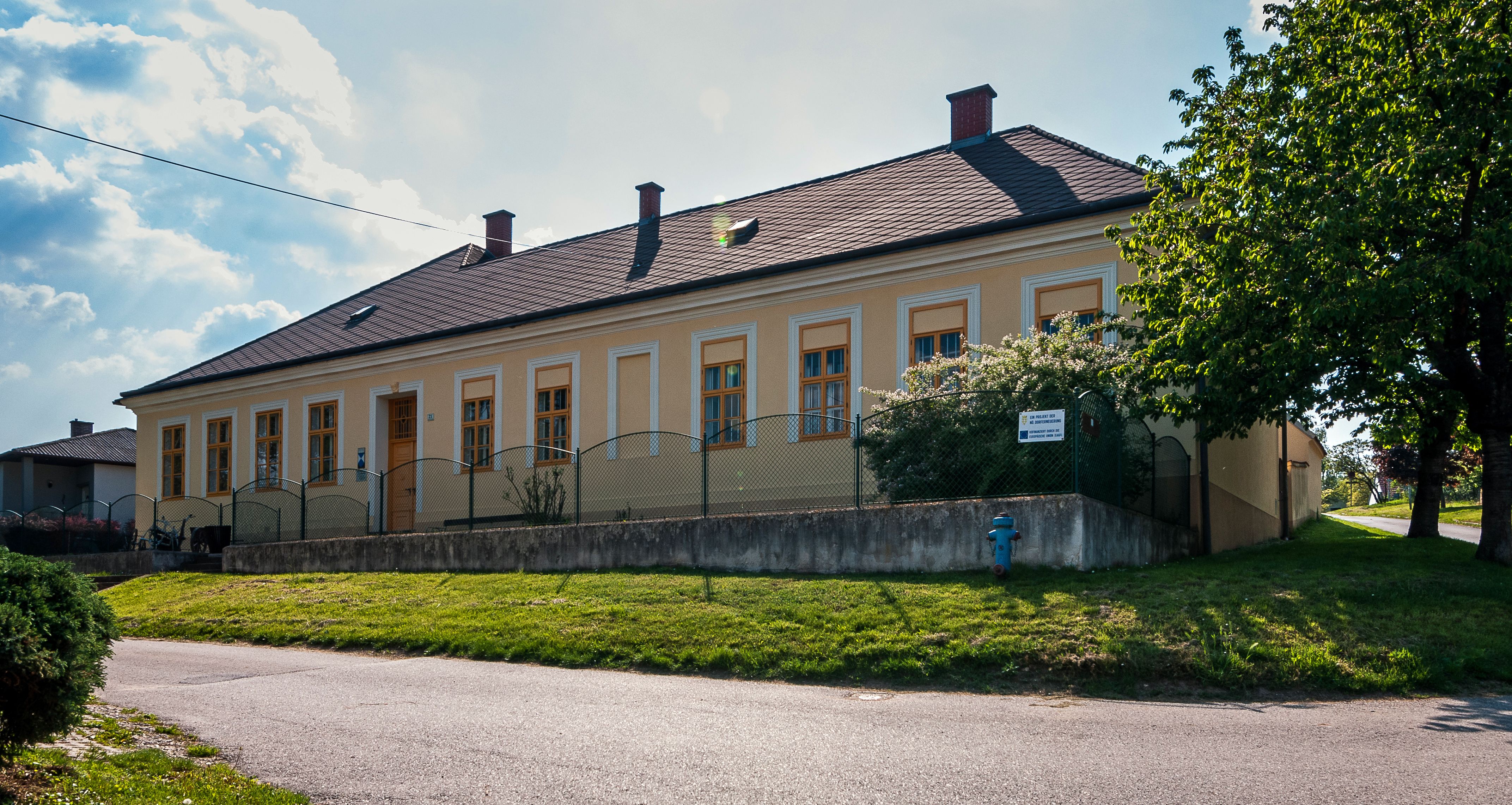 Ein gelbes Gebäude mit mehreren Fenstern und einem Zaun davor, umgeben von Bäumen und einem Rasen.