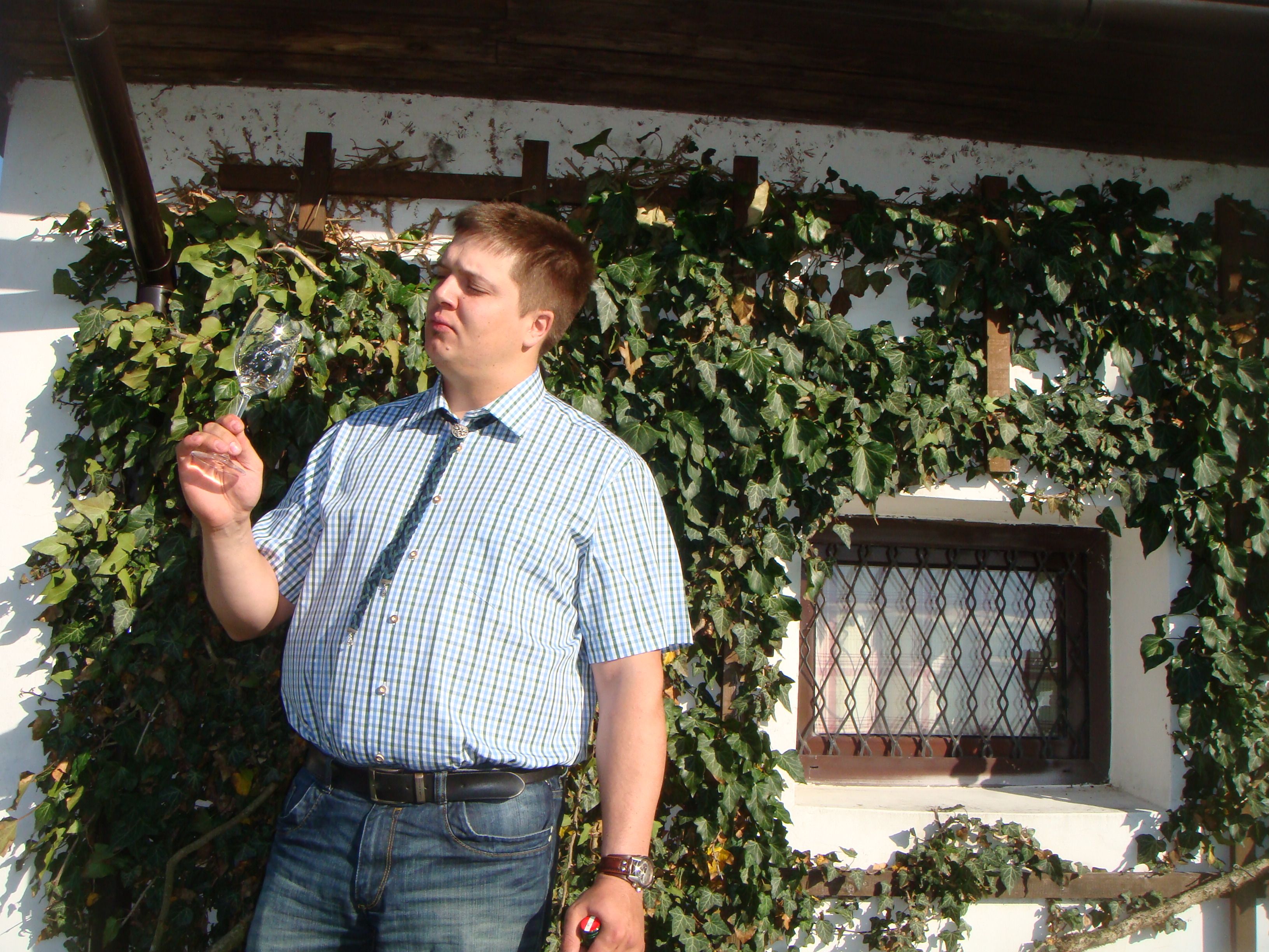 Mann in kariertem Hemd betrachtet ein Weinglas vor einer mit Efeu bewachsenen Wand.