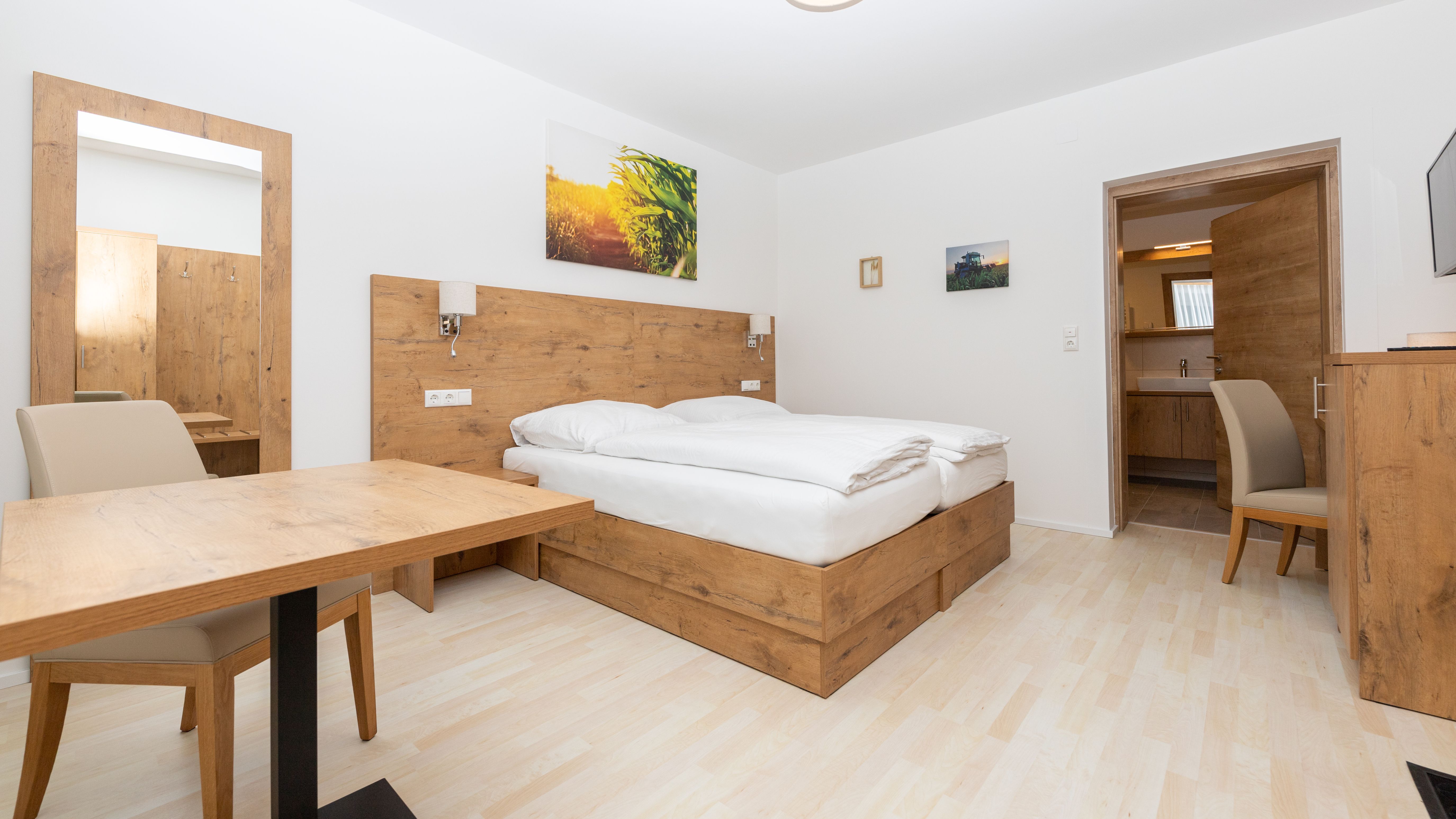 Modernes Schlafzimmer mit Holzmöbeln, Doppelbett, Tisch und Stuhl. Ein Spiegel und Bilder an der Wand, Tür führt zu einem Badezimmer.