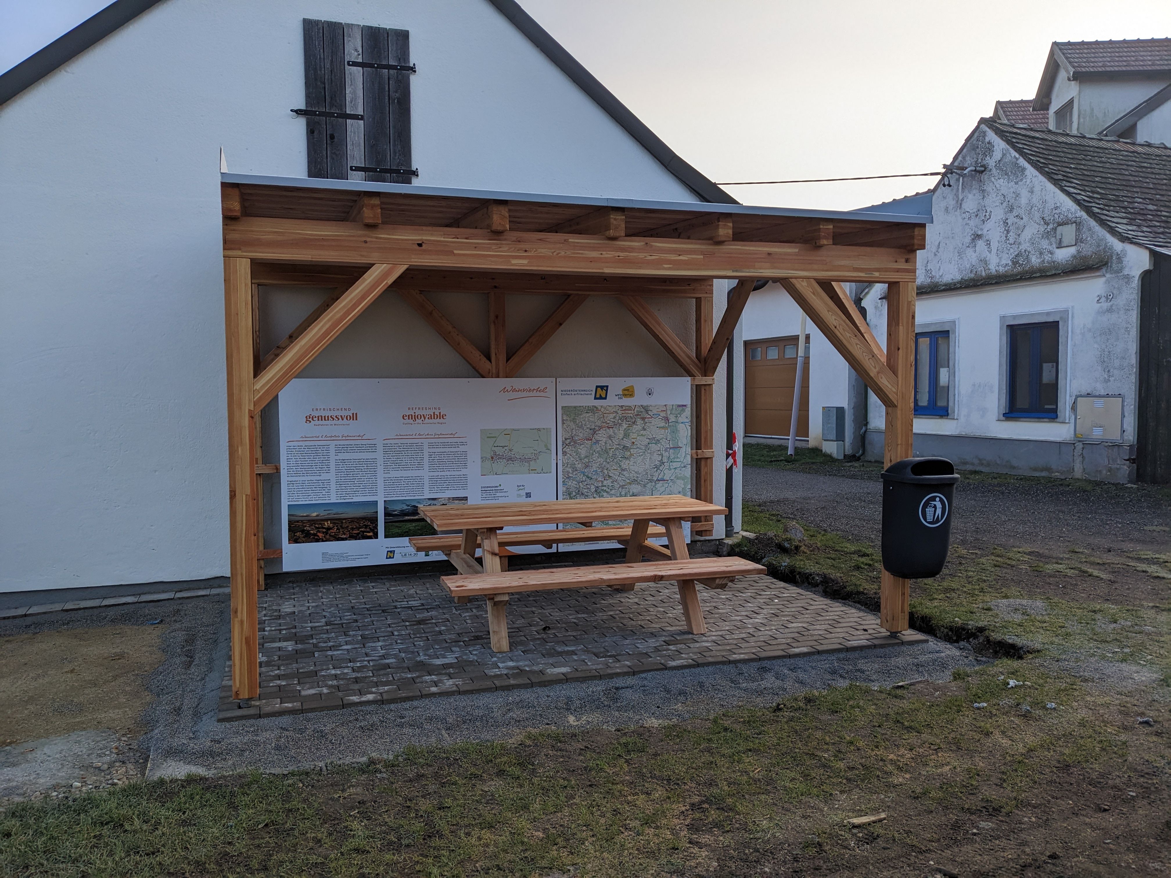 Ein Rastplatz mit Holzdach und Sitzbank vor einer weißen Hauswand, mit Informationstafeln und einem Mülleimer.
