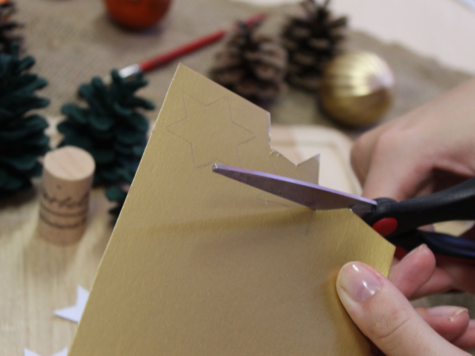 Ein Stern wird aus goldenem Papier mit einer Schere ausgeschnitten, im Hintergrund liegen Tannenzapfen, Korken und Weihnachtskugeln.