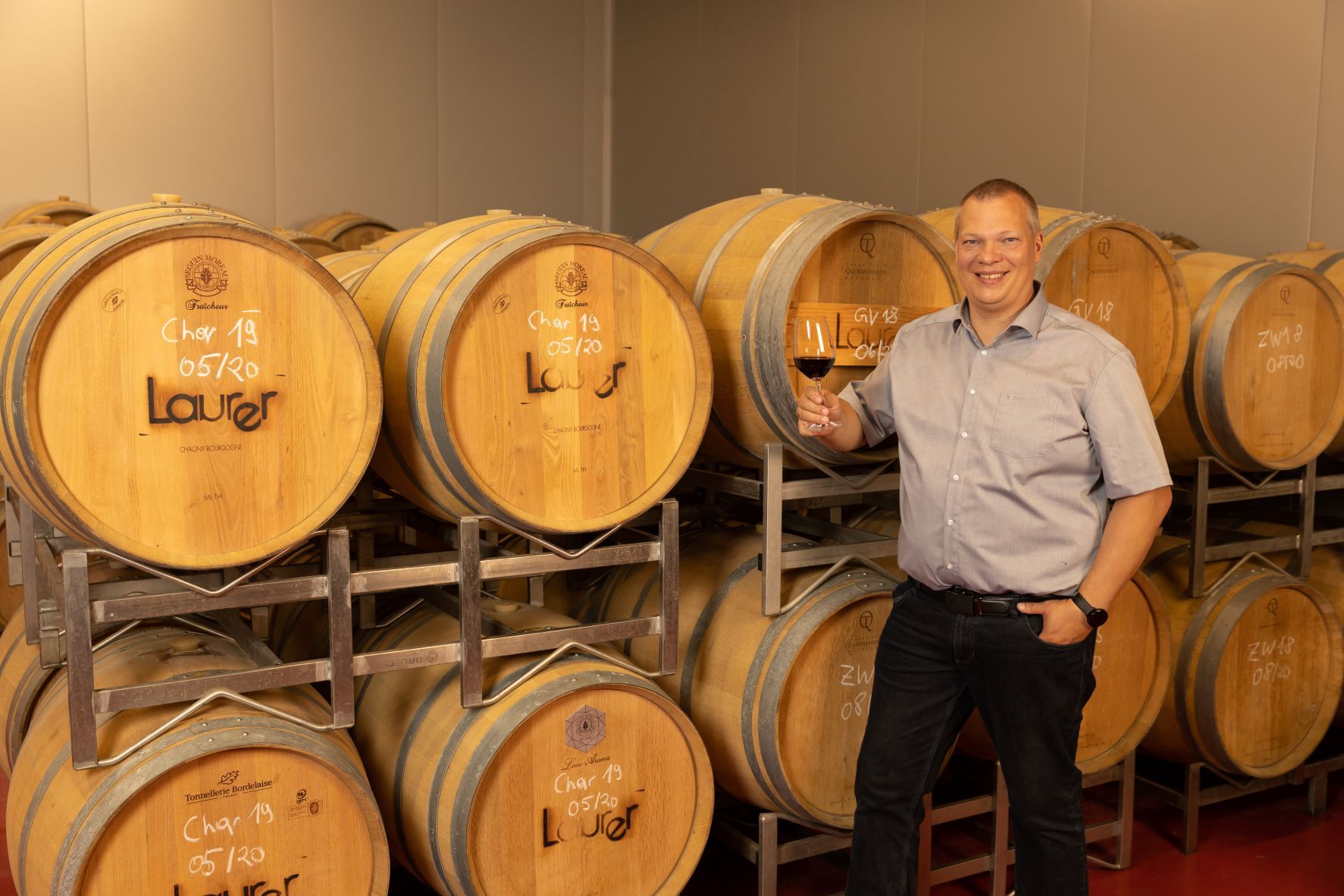 Man smiling in front of wine barrels and holding a glass of red wine.