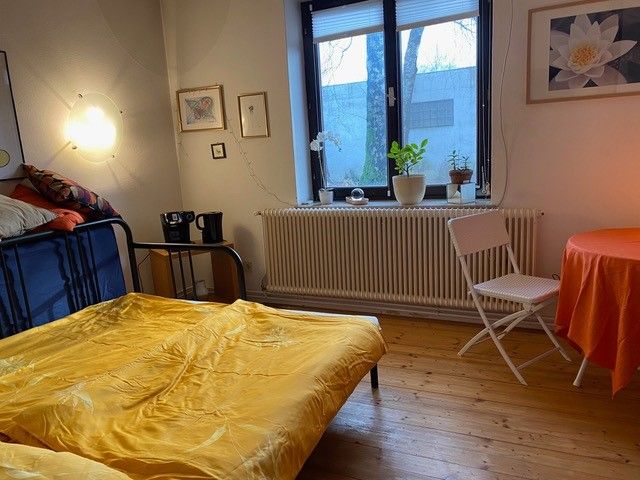 Gemütliches Schlafzimmer mit gelber Bettwäsche, Tisch mit orangener Tischdecke, Pflanzen auf Fensterbank und Wanddekorationen.