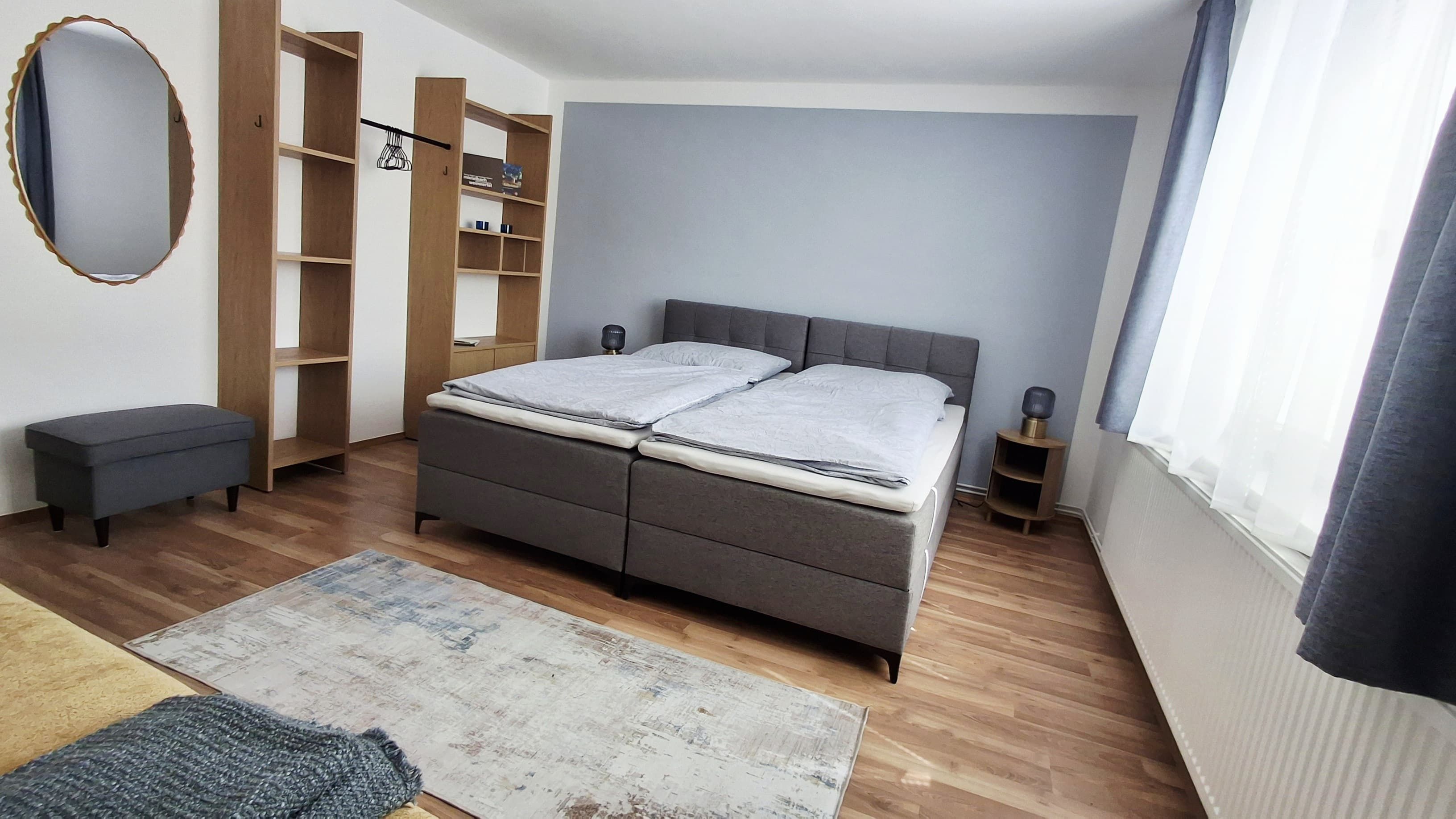 Moderná izba pre hostí s manželskou posteľou, sivými závesmi a dreveným nábytkom.