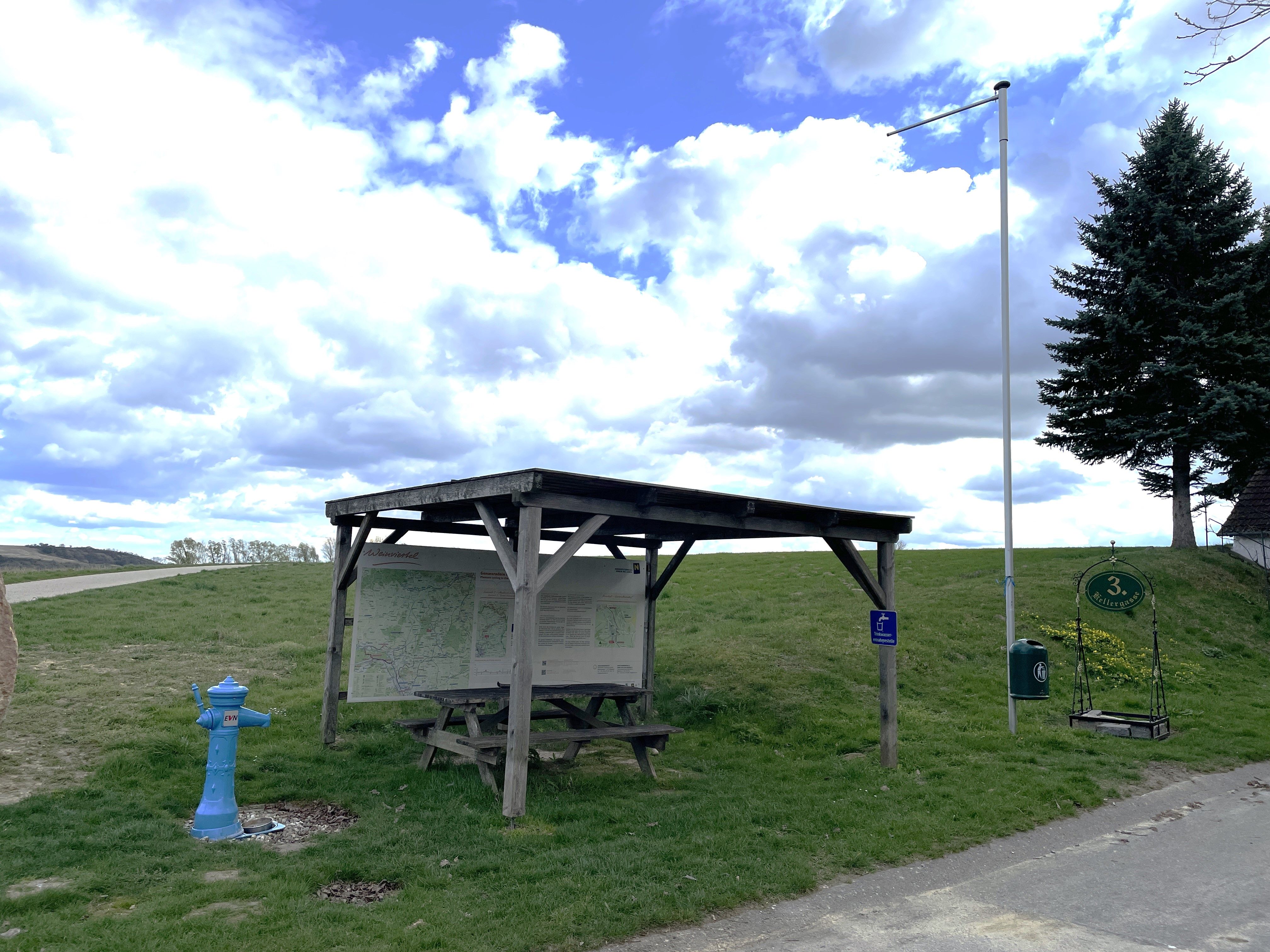 A rest area with a wooden roof, a bench, a table, and an information sign on a meadow under a cloudy sky.