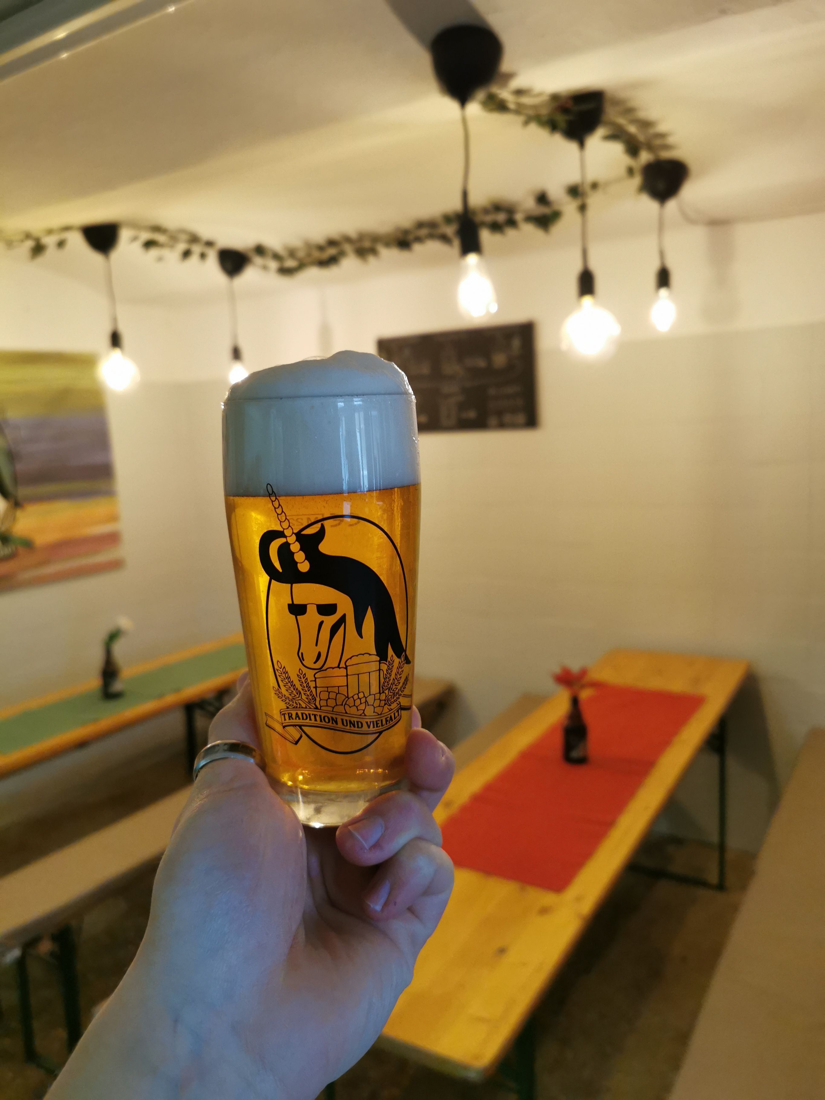 A glass of beer with a frothy head is held in a tasting room with wooden tables and fairy lights.