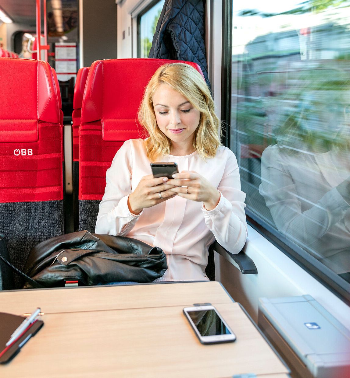 Eine junge Frau sitzt entspannt in einem modernen Zug und vertieft sich in ihr Smartphone. Die roten Sitze und das sanfte Licht schaffen eine angenehme Atmosphäre für die Reise. Draußen zieht die malerische Landschaft vorbei und verspricht Abenteuer und Entspannung.