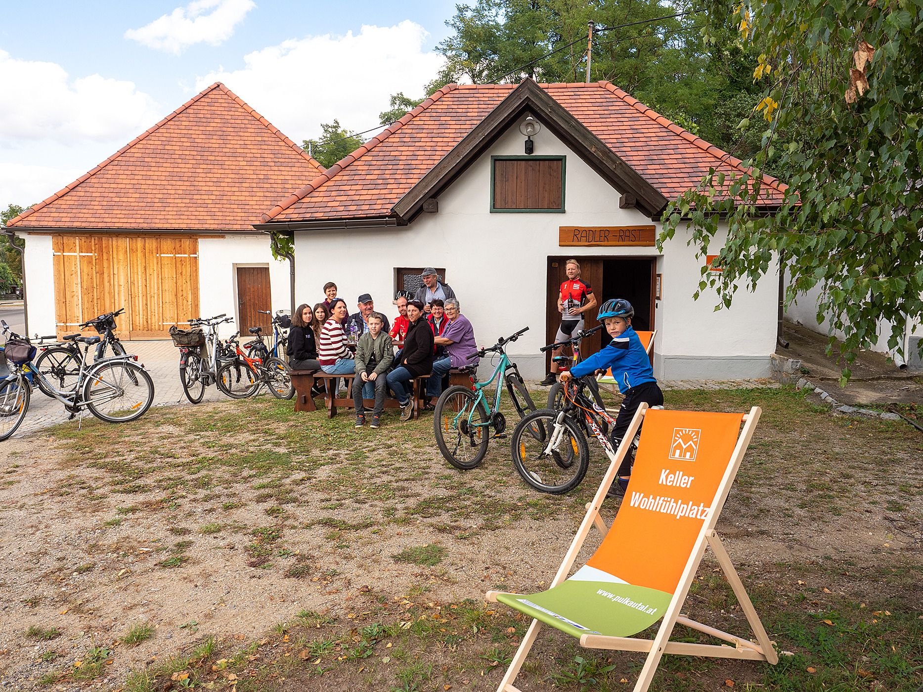 Group of people sitting in front of a building with bicycles, a child stands next to them with a bicycle. A deck chair with an inscription is in the foreground.