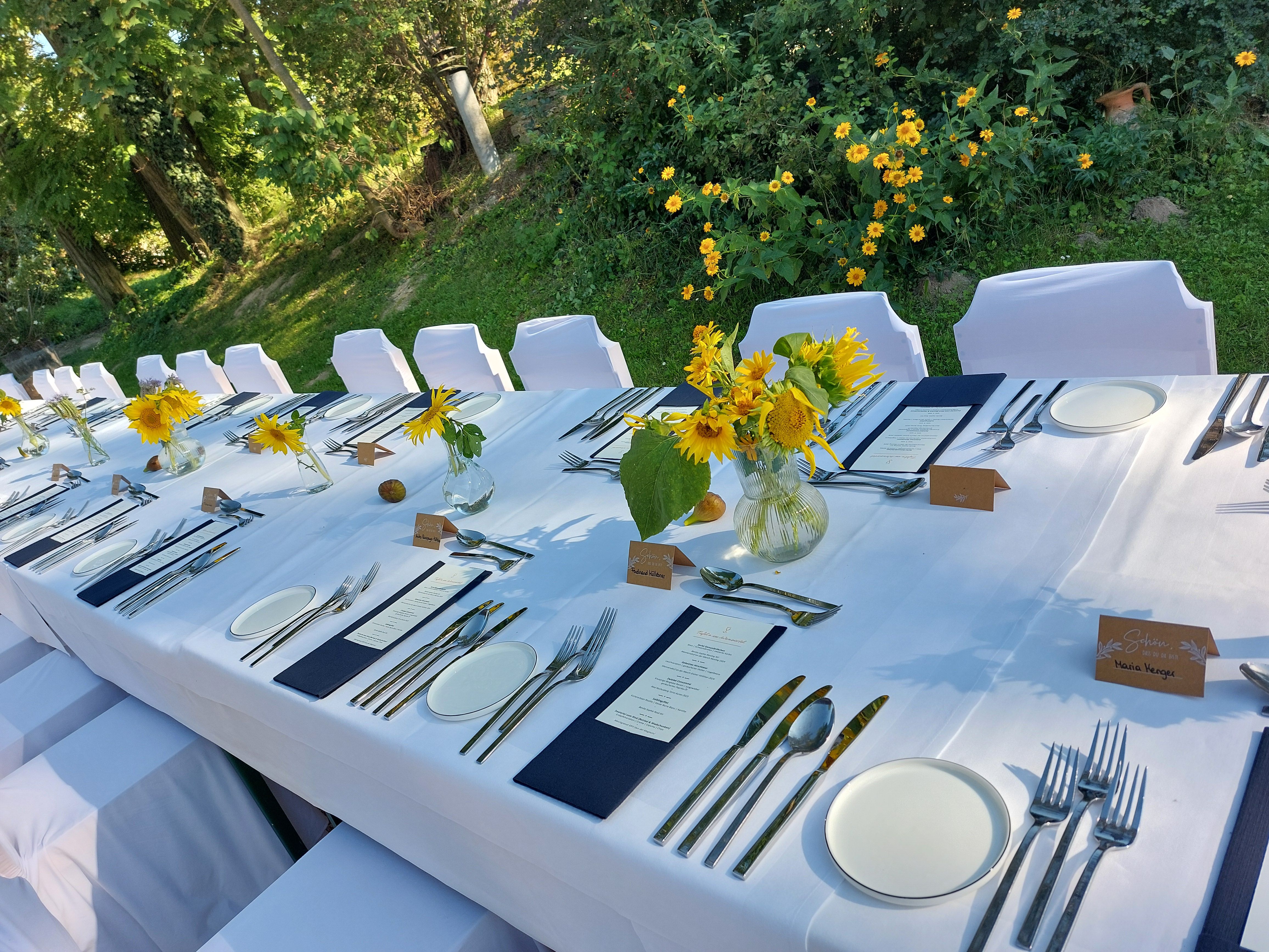 Festlich gedeckter Tisch im Freien mit weißen Tischdecken, Sonnenblumen und Besteck.