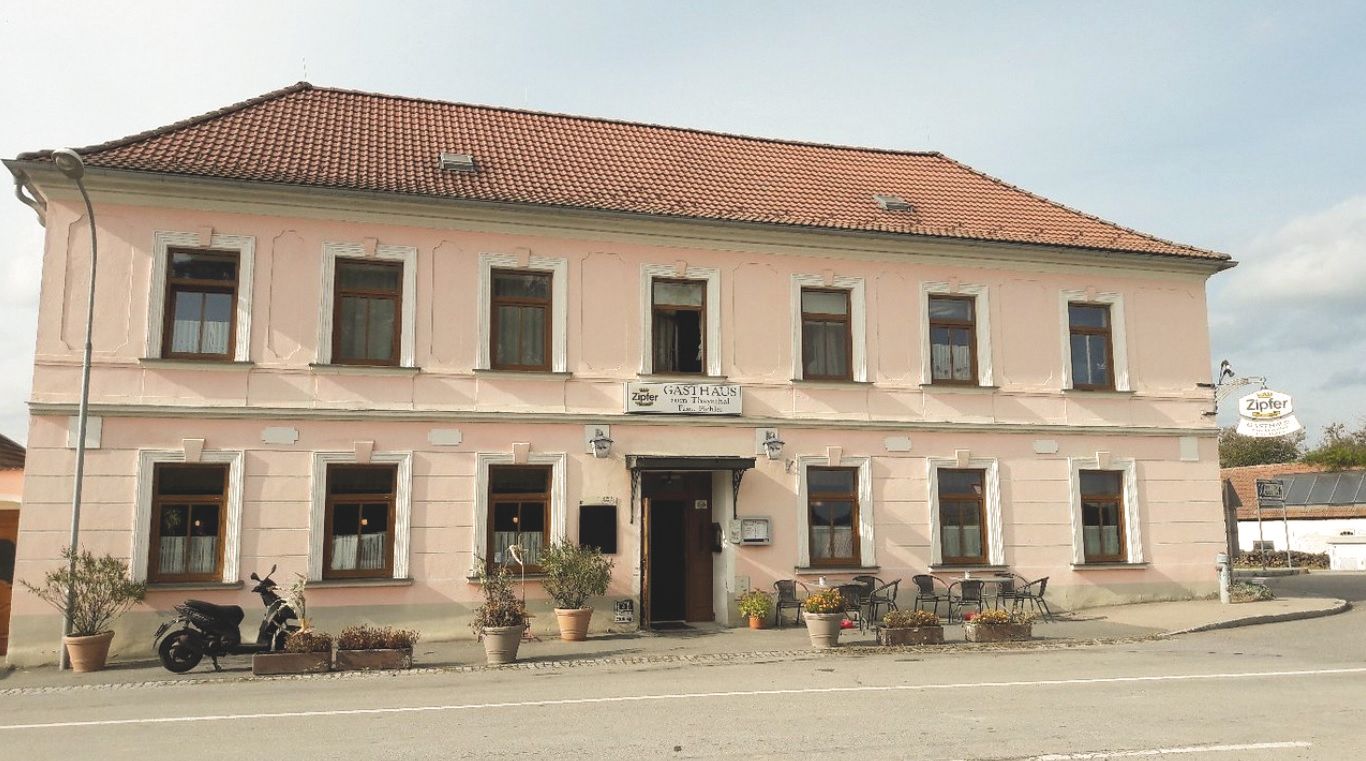 Ein zweistöckiges, rosa Gebäude mit einem roten Ziegeldach und mehreren Fenstern. Vor dem Gebäude stehen Pflanzen und ein Motorrad.