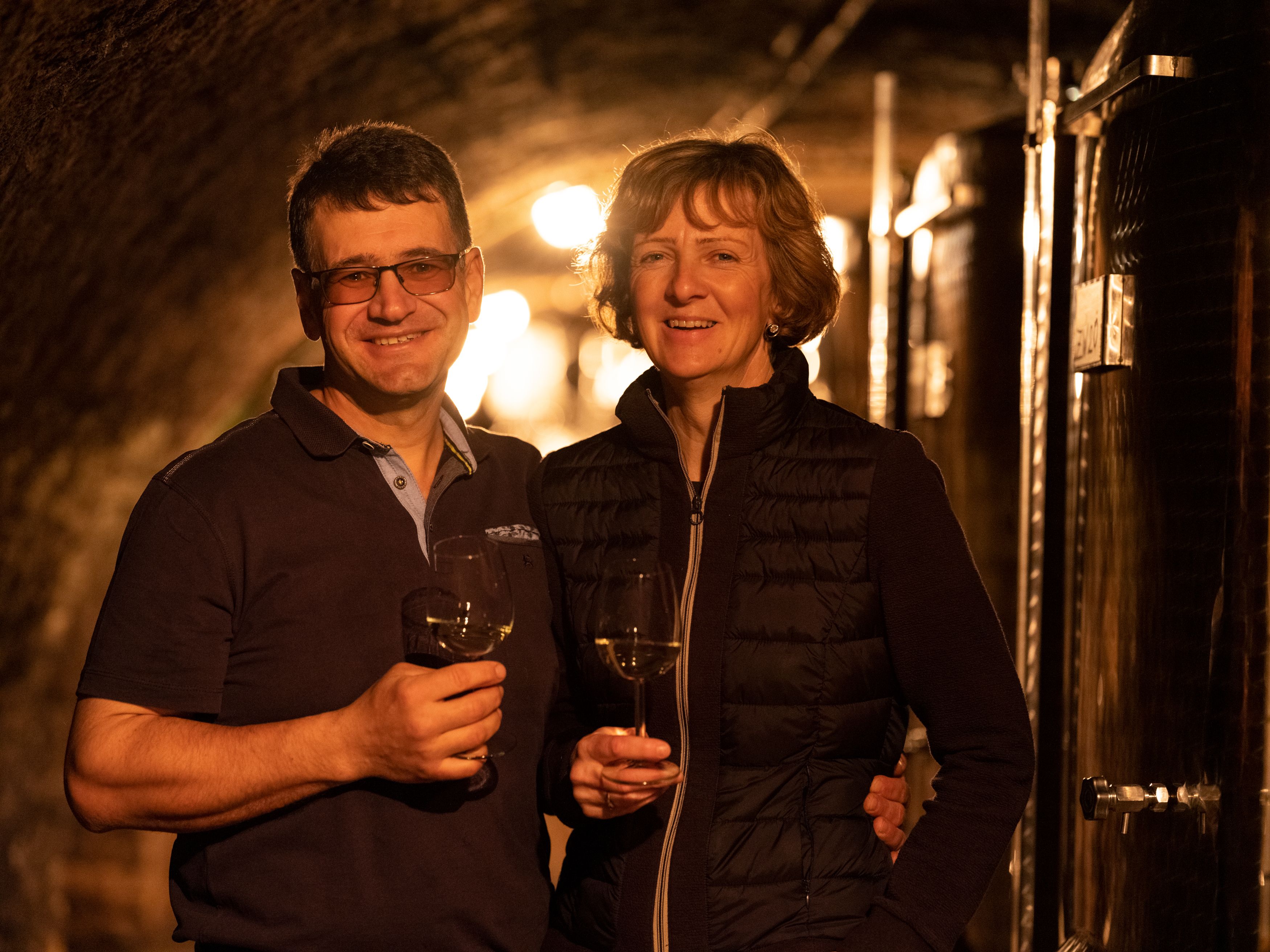 Two people in a wine cellar with wine glasses in their hands.
